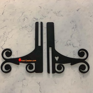 May include: Two black metal display stands with a decorative scroll design and a heart cutout. The stands are designed to hold items and have a website address printed on them.