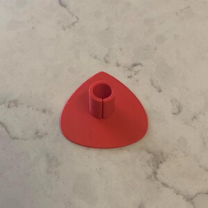 May include: A bright red, 3D printed, triangular-shaped phone stand with a round hole in the center for holding a phone.