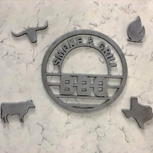 May include: A grey metal sign featuring the text "SMOKE & GRILL BBQ" in a circular design, with cut-out shapes of a longhorn, a cow, a flame, and the state of Texas. The sign is mounted on a light-coloured surface.