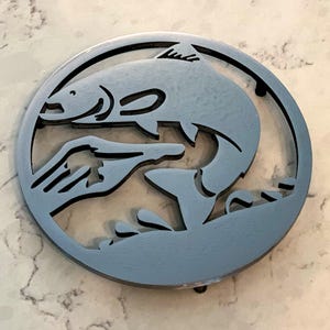 Puede incluir: Un emblema circular de metal azul claro con un pez saltando y una mano. El pez salta fuera del agua y una mano se extiende hacia él. El diseño está recortado del metal, creando un efecto de espacio negativo.