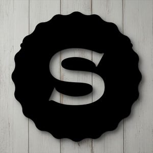 May include: Black wooden letter "S" within a decorative, scalloped-edge circle. The letter is cut out, showing the white wooden background. The design is mounted on a white wooden plank background.