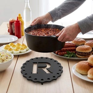 May include: A black metal trivet with a scalloped edge and the letter "R" cut out. The trivet sits on a light wood table, with a pot of chili being held above it. Other food items include devilled eggs, burgers, and hot dogs.