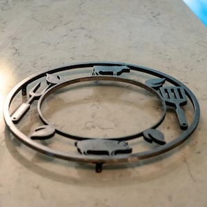 May include: A circular, metal trivet with a barbecue theme. The trivet features cut-out designs of a cow, pig, spatula, and fork, along with leaf accents. The trivet is approximately 8 inches in diameter and is designed to protect surfaces from hot items.