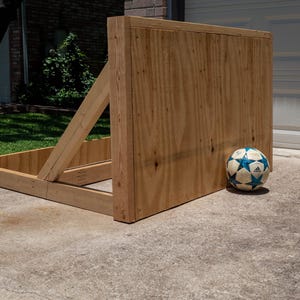 Può includere: Un rimbalzatore da calcio in legno costruito con compensato marrone chiaro e travi, dotato di un pannello rettangolare sostenuto da un supporto angolato. Un pallone da calcio con motivi bianchi e blu poggia a terra accanto alla struttura.