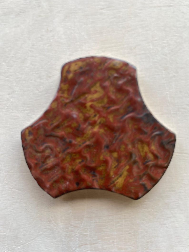 Pode incluir: Um azulejo de cer&acirc;mica vermelho e dourado com um padr&atilde;o abstrato &uacute;nico. O azulejo tem a forma de um tri&acirc;ngulo arredondado com tr&ecirc;s pontas.