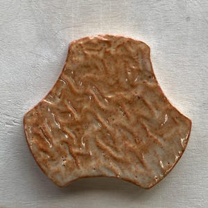 Pode incluir: Um azulejo de cer&acirc;mica de cor terracota com uma superf&iacute;cie texturizada. O azulejo tem a forma de um tri&acirc;ngulo arredondado com tr&ecirc;s lados curvos.