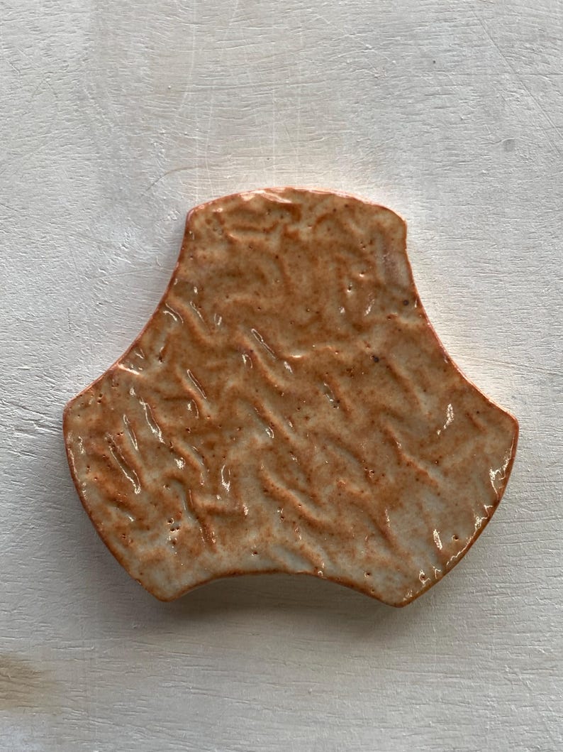 Pode incluir: Um azulejo de cer&acirc;mica de cor terracota com uma superf&iacute;cie texturizada. O azulejo tem a forma de um tri&acirc;ngulo arredondado com tr&ecirc;s lados curvos.