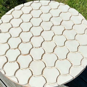 Pode incluir: Uma mesa redonda de madeira com tampo branco com um padr&atilde;o decorativo de azulejos brancos sobrepostos em forma de concha.