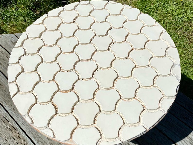 Pode incluir: Uma mesa redonda de madeira com tampo branco com um padr&atilde;o decorativo de azulejos brancos sobrepostos em forma de concha.
