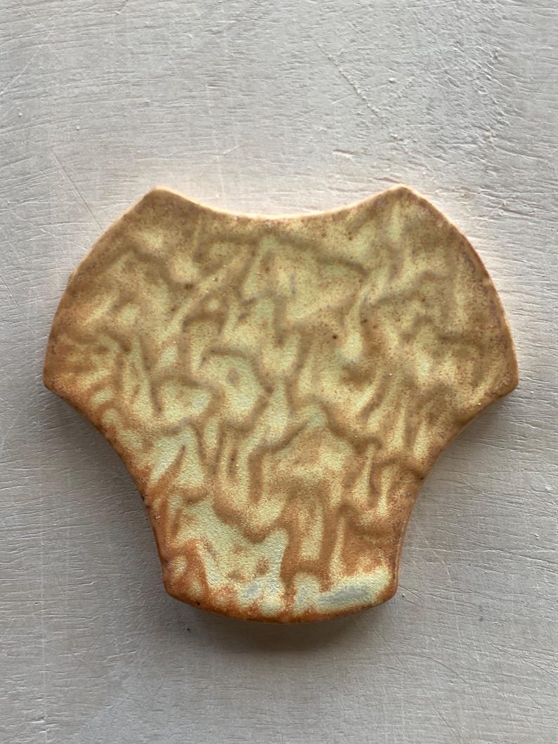 Pode incluir: Um azulejo de cer&acirc;mica com um padr&atilde;o em espiral de cor amarelo claro e marrom. O azulejo tem a forma de uma p&eacute;tala de flor com uma borda arredondada.