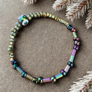 May include: Iridescent beaded bracelet with a mix of colors including green, purple, and gold. The bracelet features a large, round bead with a colorful, eye-like design. The beads are strung together to form a flexible band.
