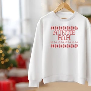 May include: White sweatshirt with red text that reads "AUNTIE FAH" and "LA LA LA LA LA LA LA LA LA". The design is in a festive, holiday style. The sweatshirt is hanging on a wooden hanger.