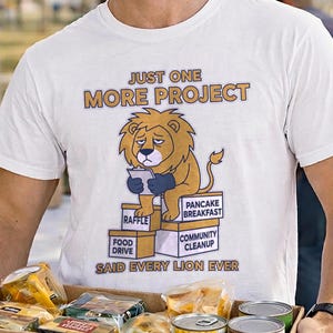 May include: A white t-shirt with the text "JUST ONE MORE PROJECT" and a cartoon lion graphic. The lion is holding a tablet, with the words "RAFFLE", "FOOD DRIVE", "PANCAKE BREAKFAST", and "COMMUNITY CLEANUP" below. A cardboard box labeled "FOOD" is being held.