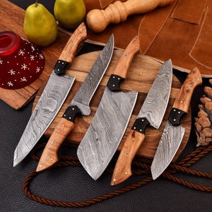 May include: A set of five kitchen knives featuring Damascus steel blades with wavy patterns, light brown wooden handles, black bolsters, and brass rivets. The knives are arranged on a wooden board alongside two green pears and a pinecone.