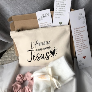 Peut inclure: Une pochette en toile blanche avec le texte "L'Amour a un nom Jesus" en lettres noires et un petit cœur rouge. La pochette est entourée de trois marque-pages avec des citations inspirantes en français. Les marque-pages sont décorés de motifs floraux. La pochette fait partie d'un coffret cadeau qui comprend également un chouchou rose et une écharpe blanche et moelleuse.