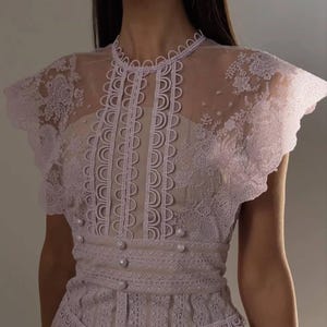 Puede incluir: Un vestido de encaje lila claro con una parte superior transparente. El vestido presenta un cuello alto, mangas cortas y una cintura ajustada con botones de perlas. Los intrincados detalles de encaje y el diseño delicado crean un look elegante y femenino.