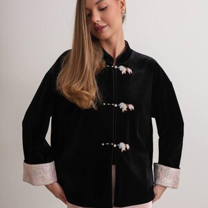 May include: Black velvet jacket with a mandarin collar and decorative closures. The jacket features iridescent cuffs and decorative closures. The jacket is a long-sleeved garment.