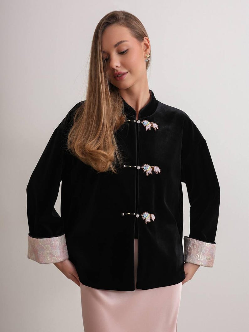 May include: Black velvet jacket with a mandarin collar and decorative closures. The jacket features iridescent cuffs and decorative closures. The jacket is a long-sleeved garment.