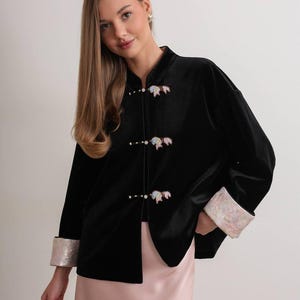 May include: Black velvet jacket with a mandarin collar and three decorative closures. The jacket features iridescent cuffs and decorative embellishments on the closures. The jacket is paired with a pink skirt.