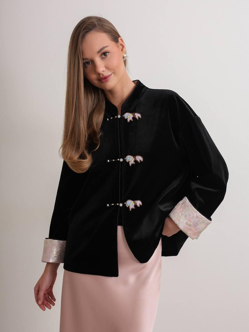 May include: Black velvet jacket with a mandarin collar and three decorative closures. The jacket features iridescent cuffs and decorative embellishments on the closures. The jacket is paired with a pink skirt.