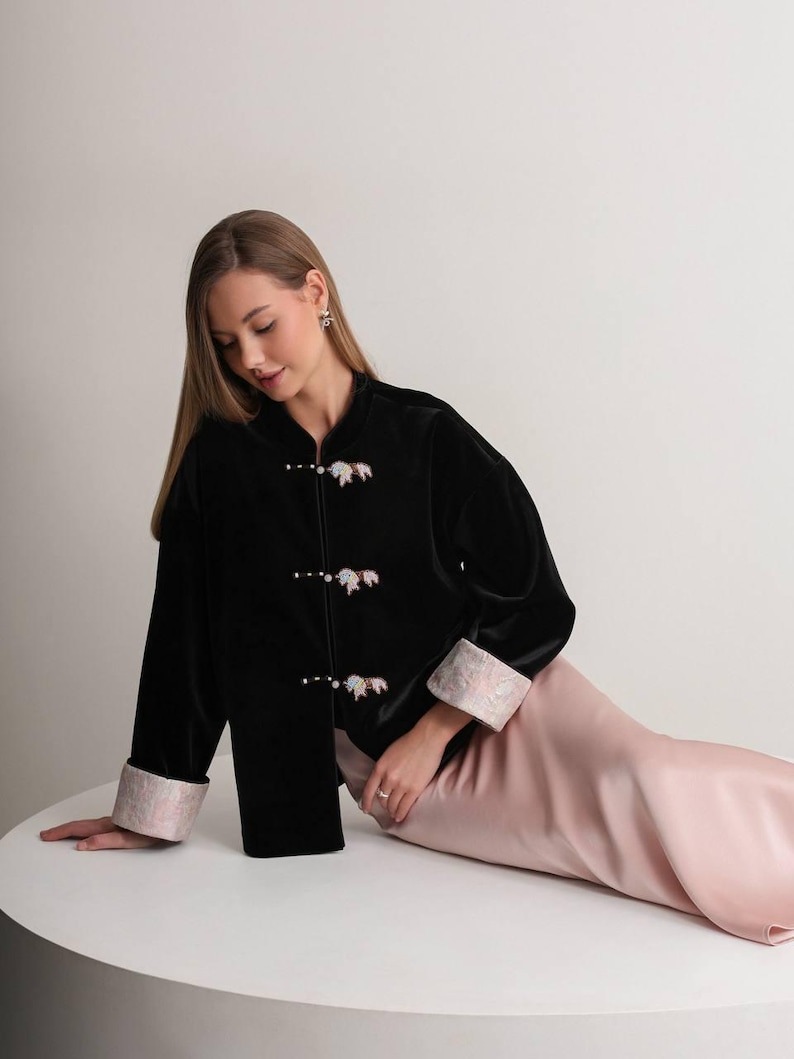 May include: A black velvet jacket with a mandarin collar and decorative closures. The jacket features light pink cuffs with a floral design. The model is wearing a light pink satin skirt.