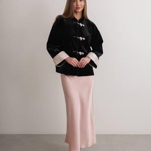 May include: A woman wearing a black velvet jacket with floral-shaped buttons and cream-colored cuffs. She is also wearing a long, light pink satin skirt and black heels. The outfit is elegant and sophisticated.