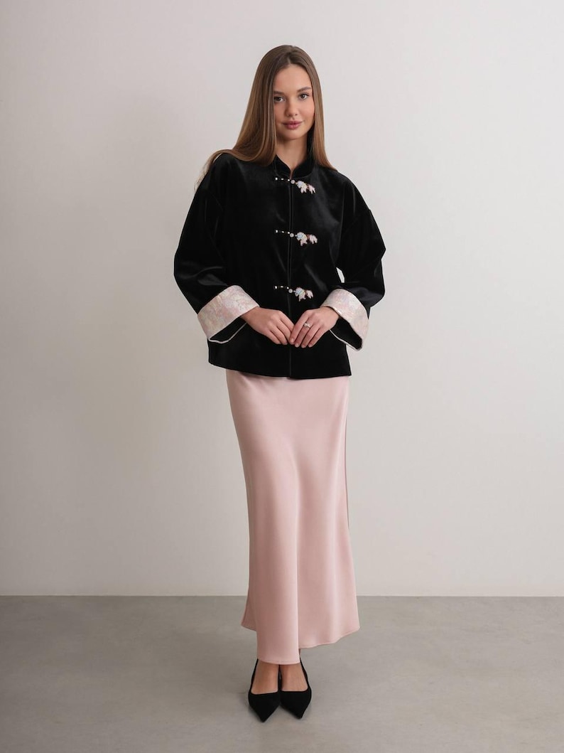May include: A woman wearing a black velvet jacket with floral-shaped buttons and cream-colored cuffs. She is also wearing a long, light pink satin skirt and black heels. The outfit is elegant and sophisticated.
