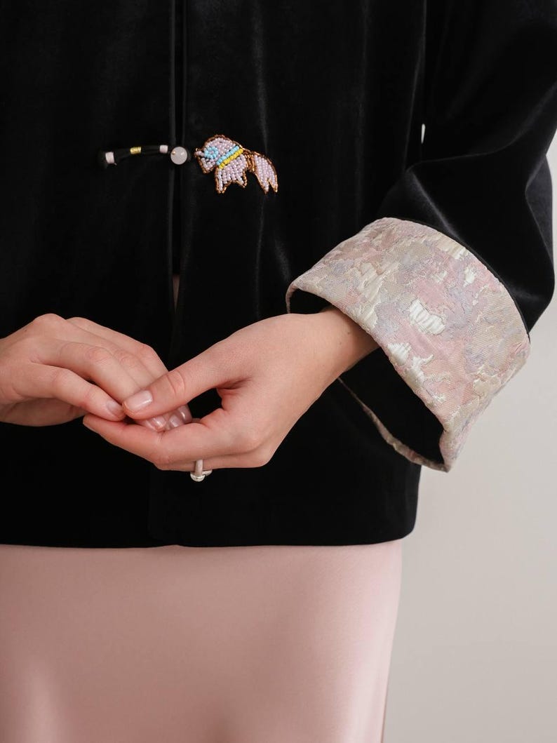 May include: Black velvet jacket with a decorative beaded pin in the shape of a small animal. The jacket features a contrasting patterned cuff in shades of pink and cream. The jacket is fastened with a toggle closure.