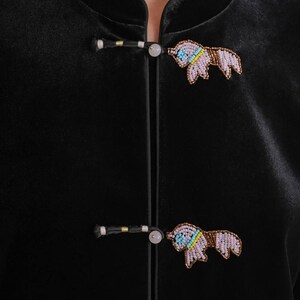May include: Close-up of a black velvet jacket with two beaded bull pins. The pins are in the shape of a bull with light purple, blue, yellow, and brown beads. The jacket has a mandarin collar and frog closures.