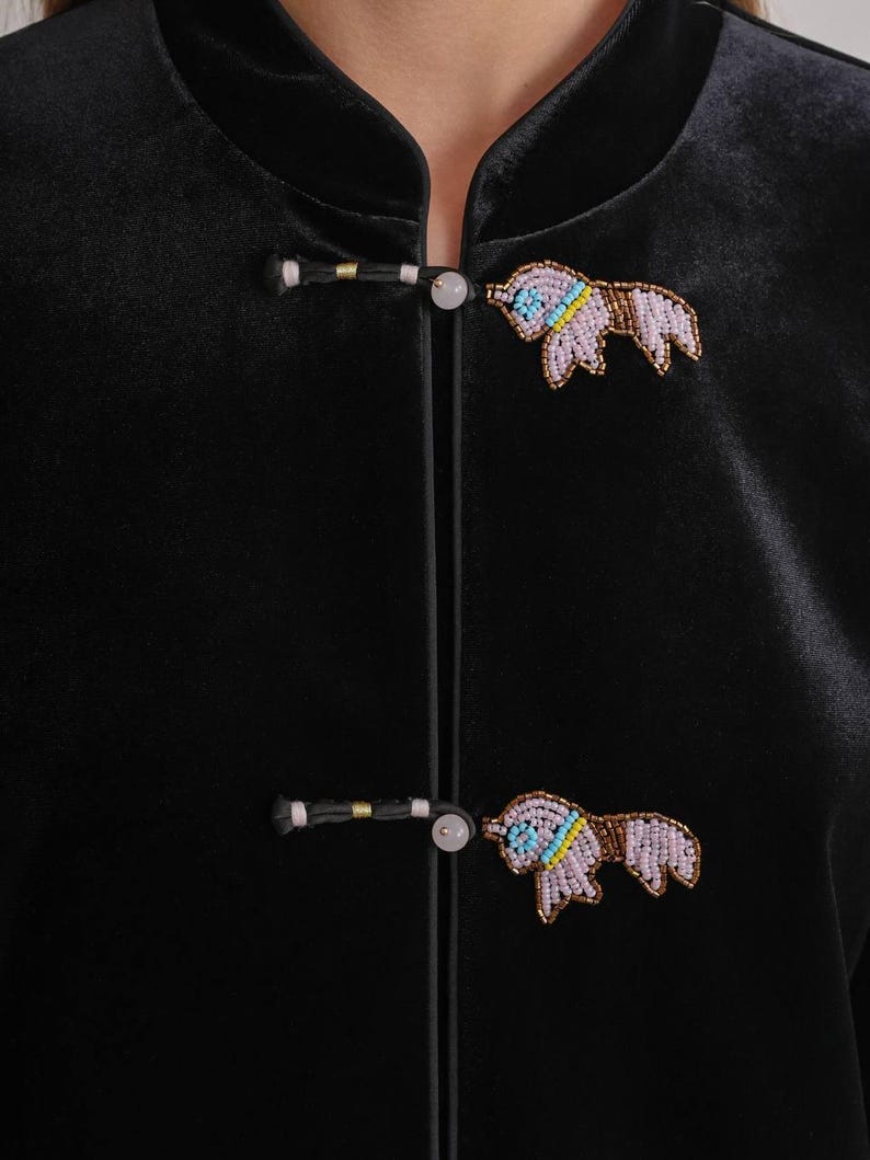 May include: Close-up of a black velvet jacket with two beaded bull pins. The pins are in the shape of a bull with light purple, blue, yellow, and brown beads. The jacket has a mandarin collar and frog closures.