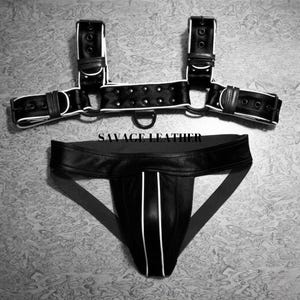 May include: Black leather harness and jockstrap set with white trim. The harness features adjustable straps and a D-ring. The jockstrap has white stripes and the text "SAVAGE LEATHER".