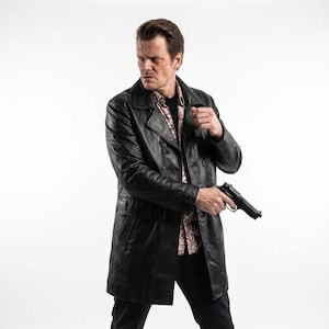 May include: A black leather trench coat with a double-breasted front, wide lapels, and two front pockets. The coat is worn over a patterned button-down shirt and dark trousers, with a black semi-automatic handgun held in the right hand.