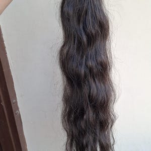 Puede incluir: Un largo mechón de extensiones de cabello castaño oscuro con una textura ondulada natural. El cabello está sujeto en la parte superior con una banda de tejido negra, mostrando un color oscuro uniforme en toda la longitud de las hebras.