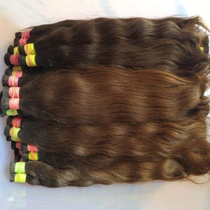 Peut inclure: Un ensemble d'extensions de cheveux bruns ondulés disposés en faisceaux horizontaux. Chaque faisceau est fixé au sommet par de petits élastiques jaune néon et rose vif, révélant un dégradé naturel de tons bruns moyens à foncés.