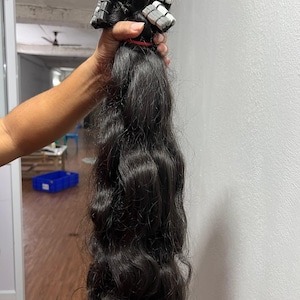 Può includere: Un fascio di extension per capelli lunghi, mossi e neri, con punti di fissaggio adesivi sulla parte superiore. I capelli presentano un motivo a onde naturale leggermente strutturato e un colore nero solido su tutta la lunghezza.