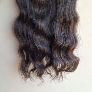 Puede incluir: Una extensión de cabello de color marrón oscuro con textura ondulada. El cabello está sujeto a una banda horizontal reforzada en la parte superior, mostrando mechones largos y fluidos con un brillo natural y un color marrón oscuro uniforme.