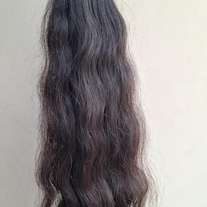 以下が含まれることがあります： 濃い茶色のウェーブのかかったヘアエクステンションの束。髪は上部で白いバンドでまとめられています。髪は自然な波模様で、根元が濃く、毛先が明るい、わずかな色のグラデーションがあります。