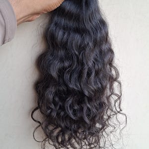 Puede incluir: Un paquete de extensiones de cabello castaño oscuro con textura ondulada. El cabello está sujeto en la parte superior con una pequeña punta negra y una banda elástica naranja claro, con mechones largos y fluidos que caen en rizos definidos.