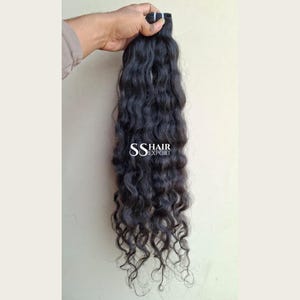 以下が含まれることがあります： ウェーブのかかった長いダークブラウンのヘアエクステンションの束。髪は上部でウェフト（横糸）によって固定されており、暗い髪の背景に対して中央に白い文字でSS HAIR EXPORTとプリントされています。