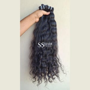 Op de afbeelding: Een bundel lange, donkere, golvende hairextensions met een weft aan de bovenkant. Het haar heeft een natuurlijke textuur en een diepzwarte kleur. De tekst SS HAIR EXPORT is in het wit in het midden van de haarbundel gedrukt.