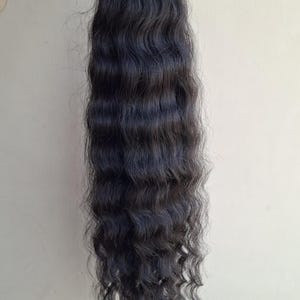 Può includere: Una lunga ciocca di extension per capelli castano scuro con una consistenza ondulata. I capelli sono fissati in alto con una trama e una fascetta in plastica bianca, mostrando un motivo a onde costante che si estende fino alle punte affusolate.