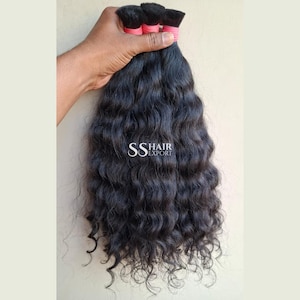 May include: A bundle of long, wavy black hair extensions secured at the top with pink bands. The hair has a natural texture and dark colour. The text SS HAIR EXPORT is printed in white in the centre of the hair bundle against the dark strands.