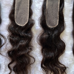Op de afbeelding: Twee donkerbruine, golvende haarvetersluitingen met een vierkante kanten basis. Het haar is gestyled in losse golven. Het kant is lichtbruin van kleur. Het haar ligt op een witte, donzige ondergrond.