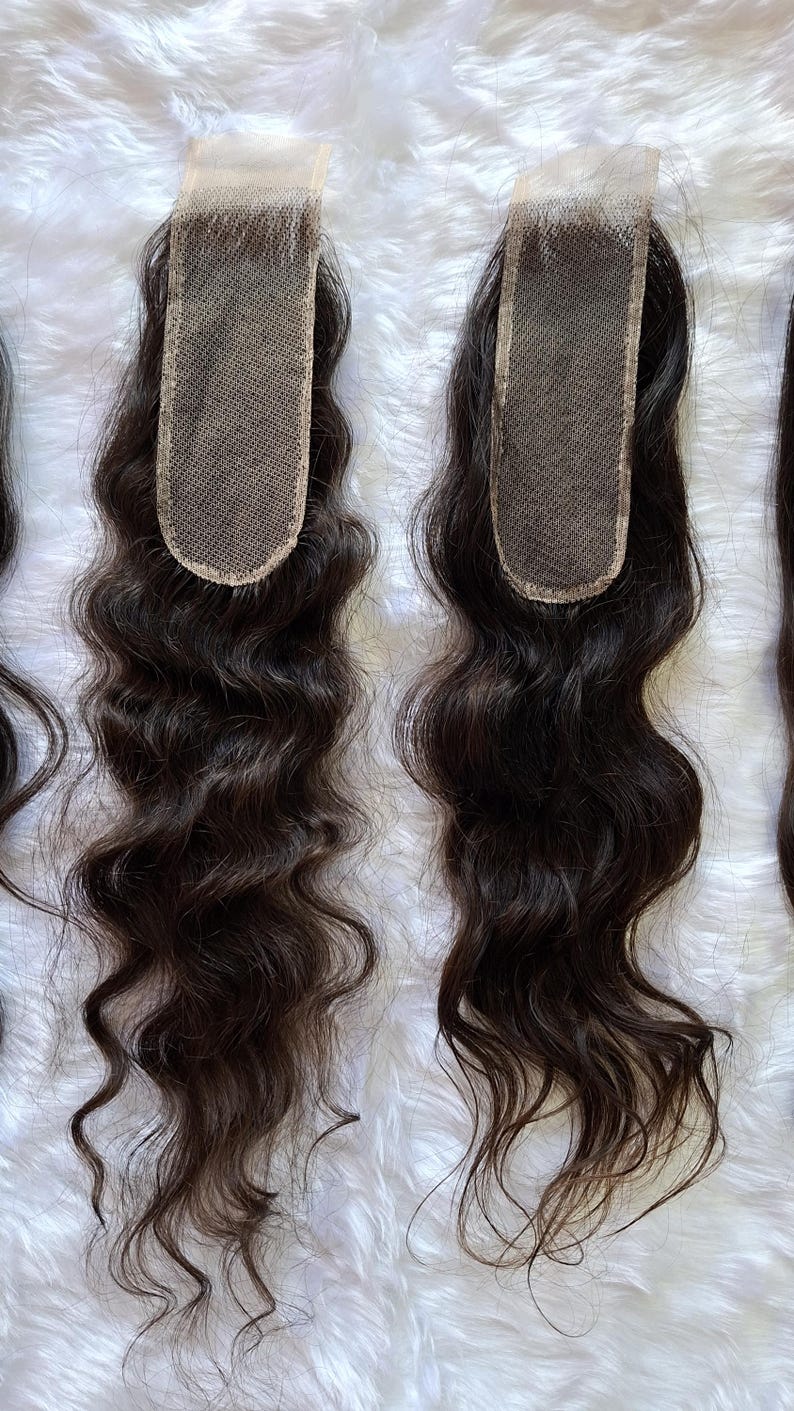 Op de afbeelding: Twee donkerbruine, golvende haarvetersluitingen met een vierkante kanten basis. Het haar is gestyled in losse golven. Het kant is lichtbruin van kleur. Het haar ligt op een witte, donzige ondergrond.
