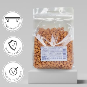 以下が含まれることがあります： 茶色のタイガーナッツが入った透明なプラスチック製のスタンドパック。ラベルにはTiger Nutsの文字、バーコード、QRコードが記載されています。左側の3つの円形アイコンは、Long Shelf Life、Clean & Sorted、Premium Tigernutsという特徴を強調しています。