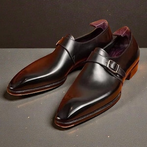 K&ouml;nnte beinhalten: Ein Paar schwarze Lederschuhe mit gl&auml;nzendem Finish. Die Schuhe haben eine spitze Zehenpartie, einen Schnallenverschluss und einen niedrigen Absatz. Die Innenseite des Schuhs ist mit einem lila Stoff gef&uuml;ttert. Die Schuhe werden auf einer dunklen Oberfl&auml;che pr&auml;sentiert.