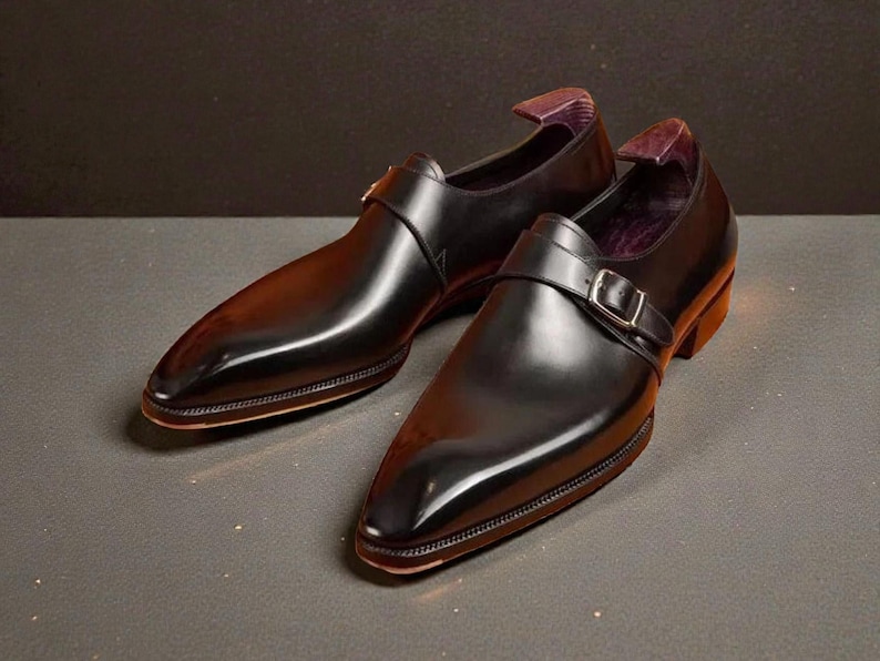 K&ouml;nnte beinhalten: Ein Paar schwarze Lederschuhe mit gl&auml;nzendem Finish. Die Schuhe haben eine spitze Zehenpartie, einen Schnallenverschluss und einen niedrigen Absatz. Die Innenseite des Schuhs ist mit einem lila Stoff gef&uuml;ttert. Die Schuhe werden auf einer dunklen Oberfl&auml;che pr&auml;sentiert.