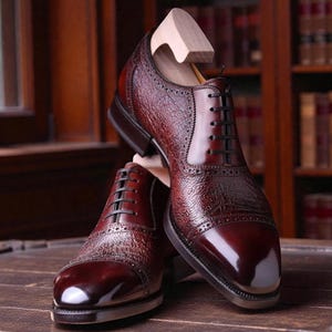 Puede incluir: Un par de zapatos de vestir de cuero burdeos con un acabado texturizado y puntera pulida. Los zapatos tienen cordones negros y se exhiben con hormas de madera. El fondo incluye una superficie de madera y una estanter&iacute;a.