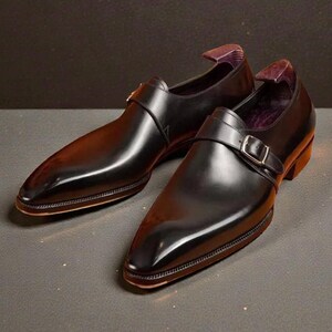 K&ouml;nnte beinhalten: Ein Paar schwarze Lederschuhe mit gl&auml;nzender Oberfl&auml;che. Die Schuhe haben eine spitze Zehenpartie, einen Schnallenverschluss und einen niedrigen Absatz. Die Innenseite des Schuhs ist mit einem lila Stoff gef&uuml;ttert. Die Schuhe werden auf einer dunklen Oberfl&auml;che pr&auml;sentiert.