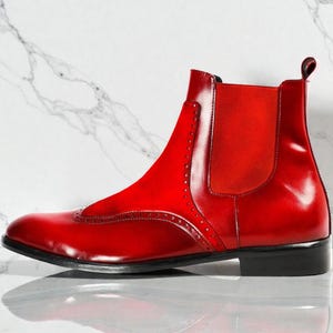 May include: A single, glossy red Chelsea boot with a black sole. The boot features a brogue design on the toe and a red elastic panel on the side. The boot is set against a white marble background.